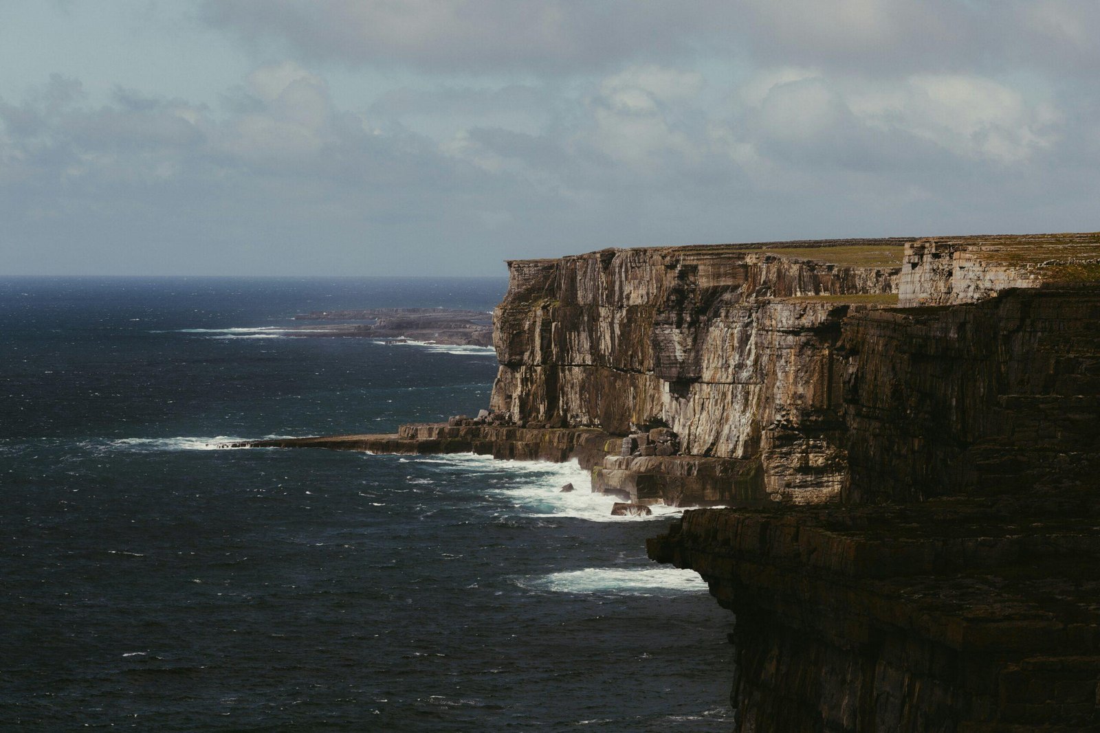 Dramatic cliffs meet the turbulent ocean under a cloudy sky.