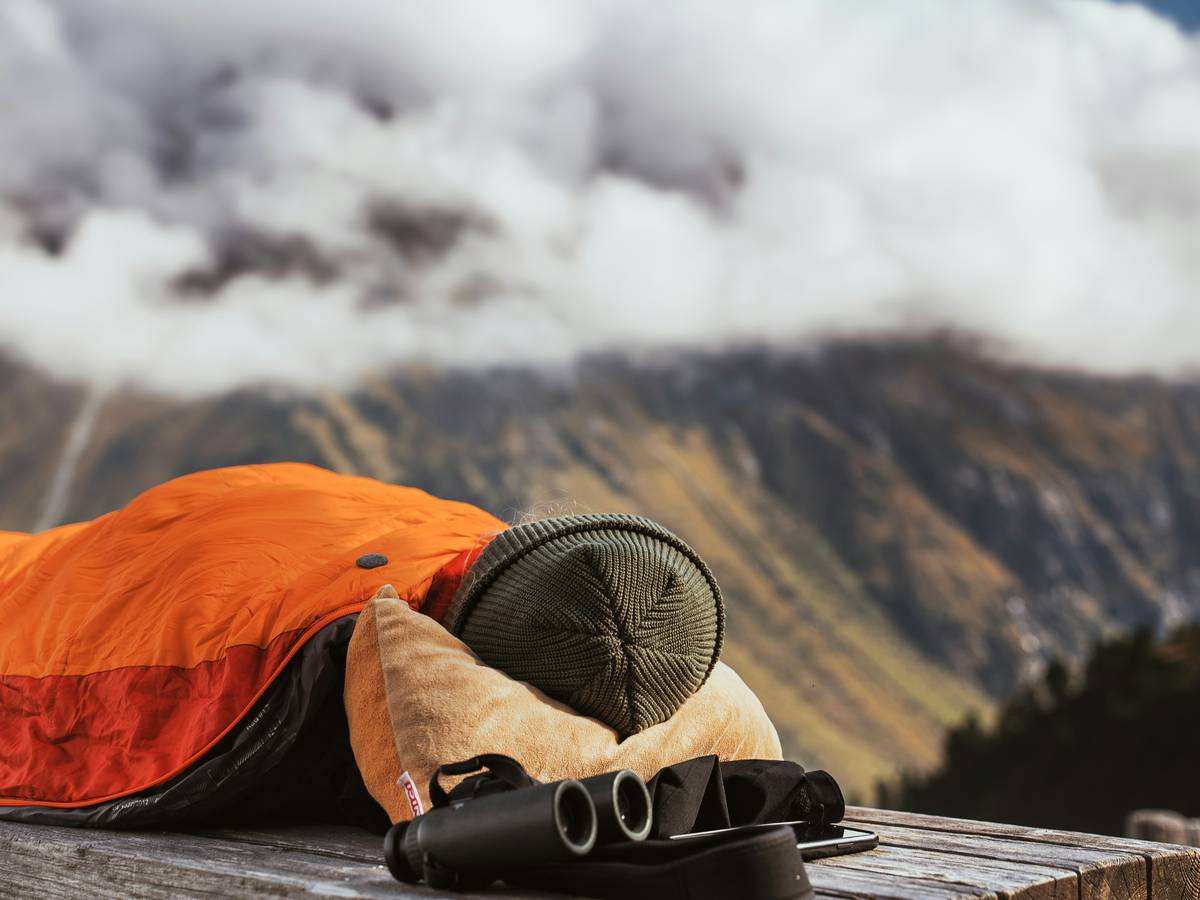 A person in an orange sleeping bag, lying on a wooden surface outdoors, likely in a mountainous area. Binoculars are nearby, and the foggy mountain backdrop creates a peaceful, remote, and reflective atmosphere.