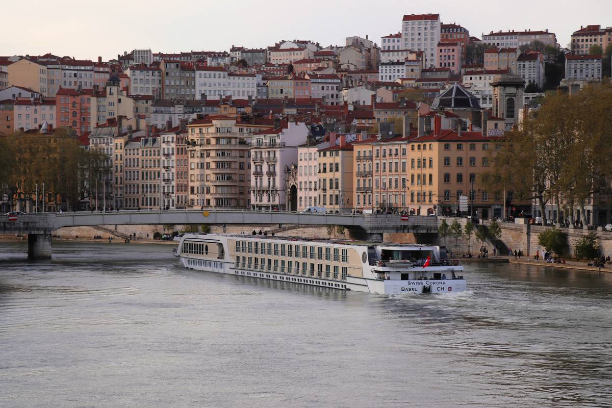 A long river cruise ship sails along the Saône in Lyon, passing pastel riverfront facades with the stacked hillside apartments of Croix-Rousse rising behind—calm water, soft evening light, classic citylife by the river.
